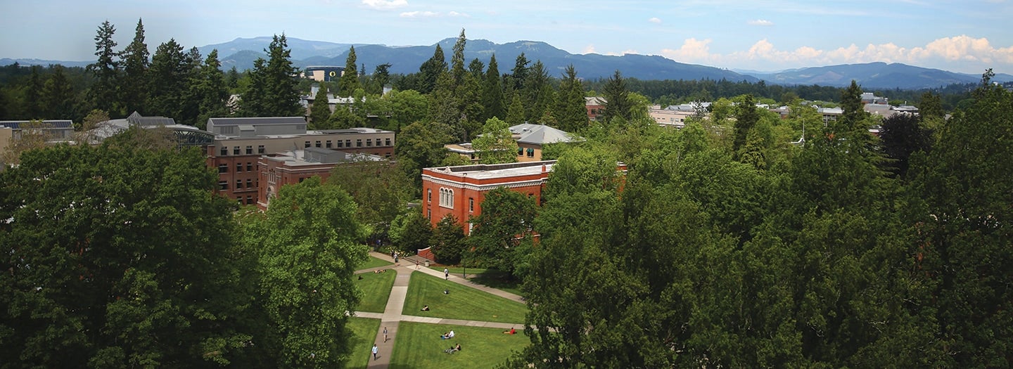 From the top of a tall building, looking over the UO campus quadrangle toward Lillis College of Business complex, the "O" logo on Autzen Stadium, and the Coburg Hills rising in the distance.