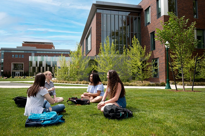 Four students sitting together on a lawn in front of the EMU.